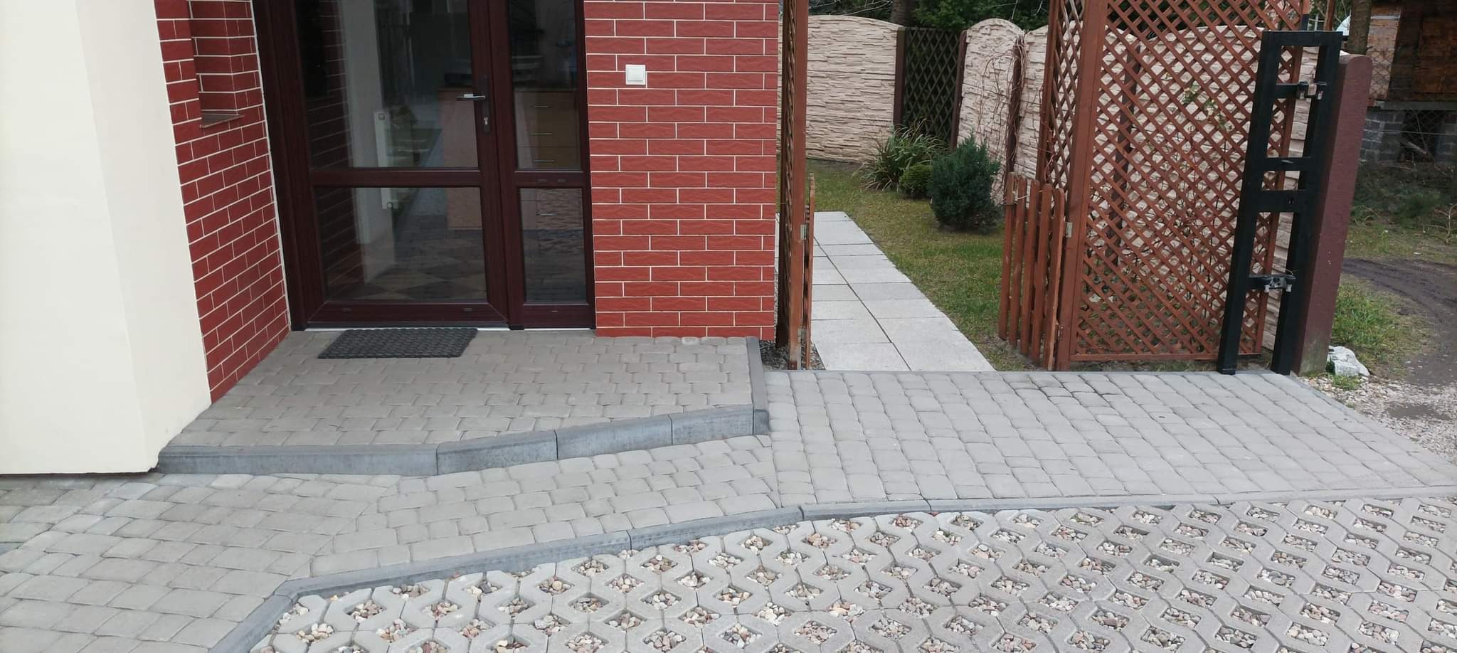 Ułożona kostka brukowa przed domem w Rokietnicy, z widocznym wejściem do budynku, schodami z kostki oraz ażurową nawierzchnią z kamieniami, prowadząca do drewnianej furtki i ścieżki w ogrodzie.