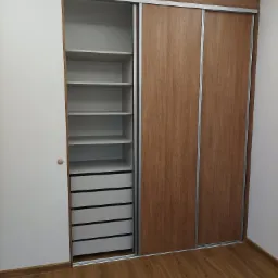 Wnętrze szafy przesuwnej z drewnianymi drzwiami i białymi półkami oraz szufladami, widoczne na tle drewnianej podłogi.