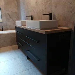 Nowoczesna łazienka z podwójną umywalką nablatową, czarną szafką z szufladami i bateriami w tym samym kolorze, na tle ściany wyłożonej kamiennymi płytkami.