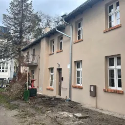 Odnowiona elewacja budynku z beżowym tynkiem, nowymi oknami z białymi ramami i ceglanymi parapetami, widoczne nowe rynny i fragment dachu, na pierwszym planie teren po pracach wykończeniowych.