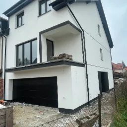 Nowoczesny dom jednorodzinny z białą elewacją i czarnymi akcentami, widok z dołu, teren wokół domu w trakcie prac wykończeniowych, widoczne elementy ogrodzenia i porozrzucane materiały budowlane.