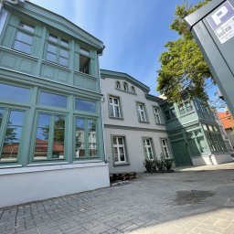 USŁUGI OGÓLNOBUDOWLANE DAWID WRYCZA - Odnowiona elewacja budynku z zielonymi oknami, widok z dołu na tle błękitnego nieba, kostka brukowa na pierwszym planie, słupek parkingowy po prawej.