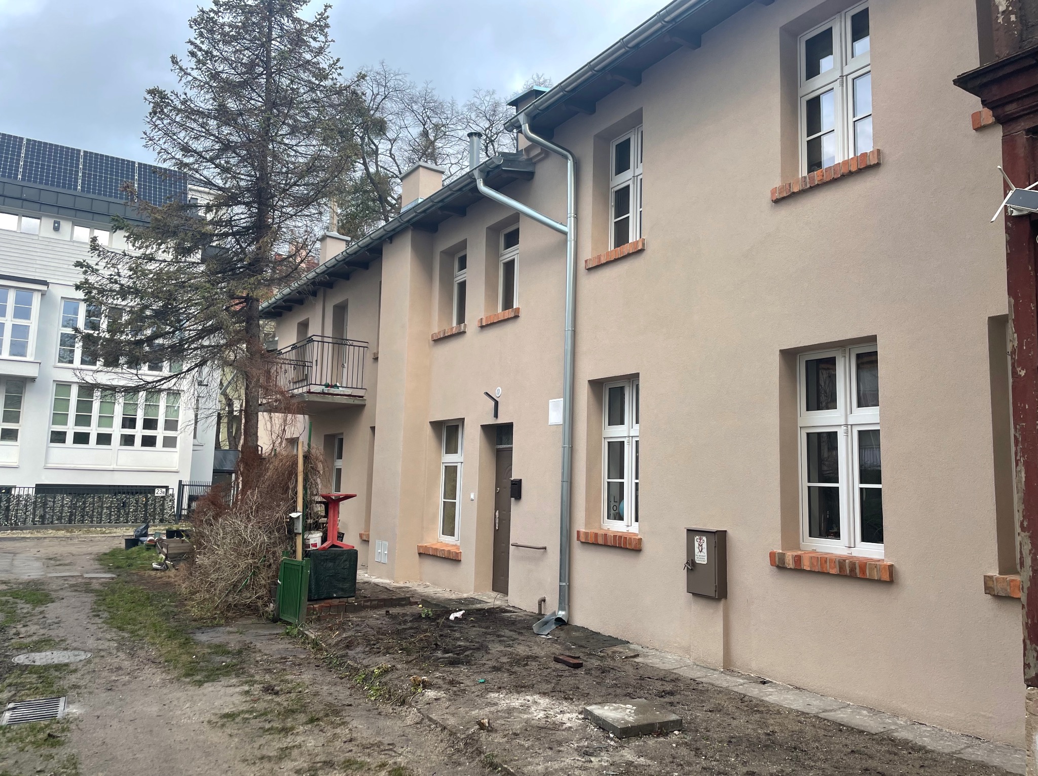 Odnowiona elewacja budynku z beżowym tynkiem, nowymi oknami z białymi ramami i ceglanymi parapetami, widoczne nowe rynny i fragment dachu, na pierwszym planie teren po pracach wykończeniowych.