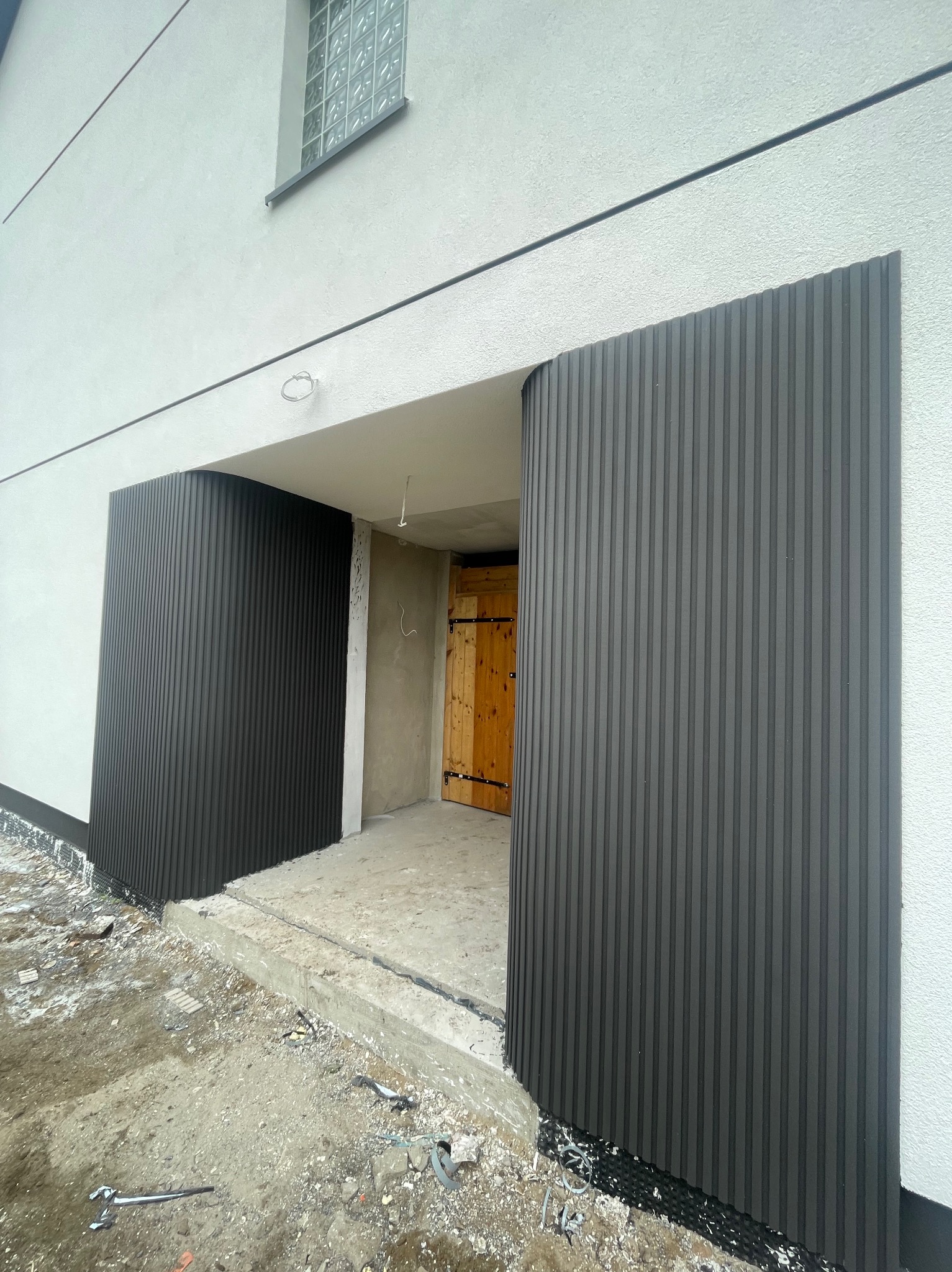 Montaż paneli elewacyjnych w kolorze antracytowym przy wejściu do budynku, widoczne narzędzia i niezagospodarowany teren.