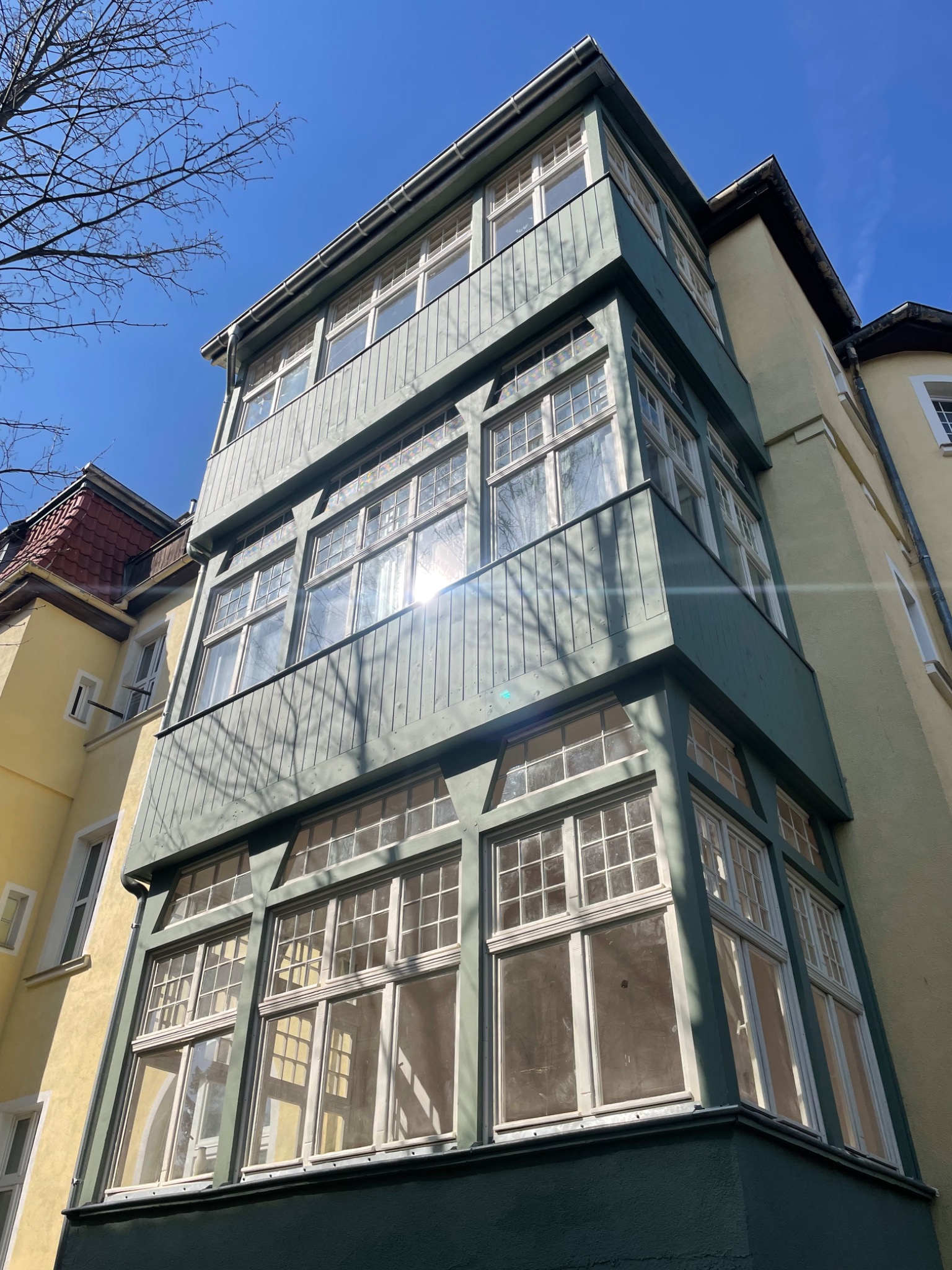 Niski kąt widzenia na odnowioną, trzypiętrową część budynku z licznymi oknami w białych ramach, pomalowaną na zielono i żółto, z widocznym fragmentem czerwonego dachu sąsiedniego budynku...