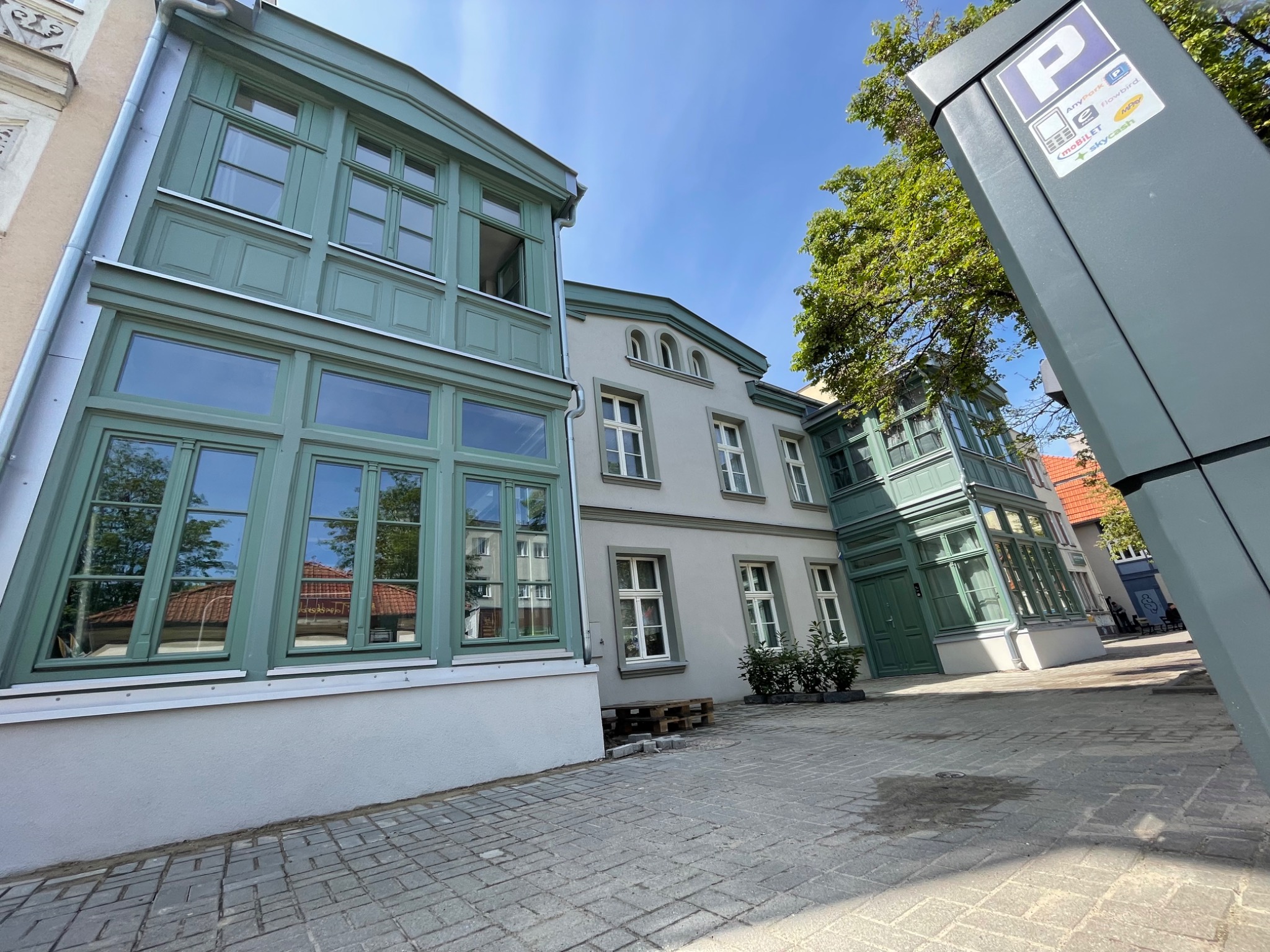 Odnowiona elewacja budynku z zielonymi oknami, widok z dołu na tle błękitnego nieba, kostka brukowa na pierwszym planie, słupek parkingowy po prawej.