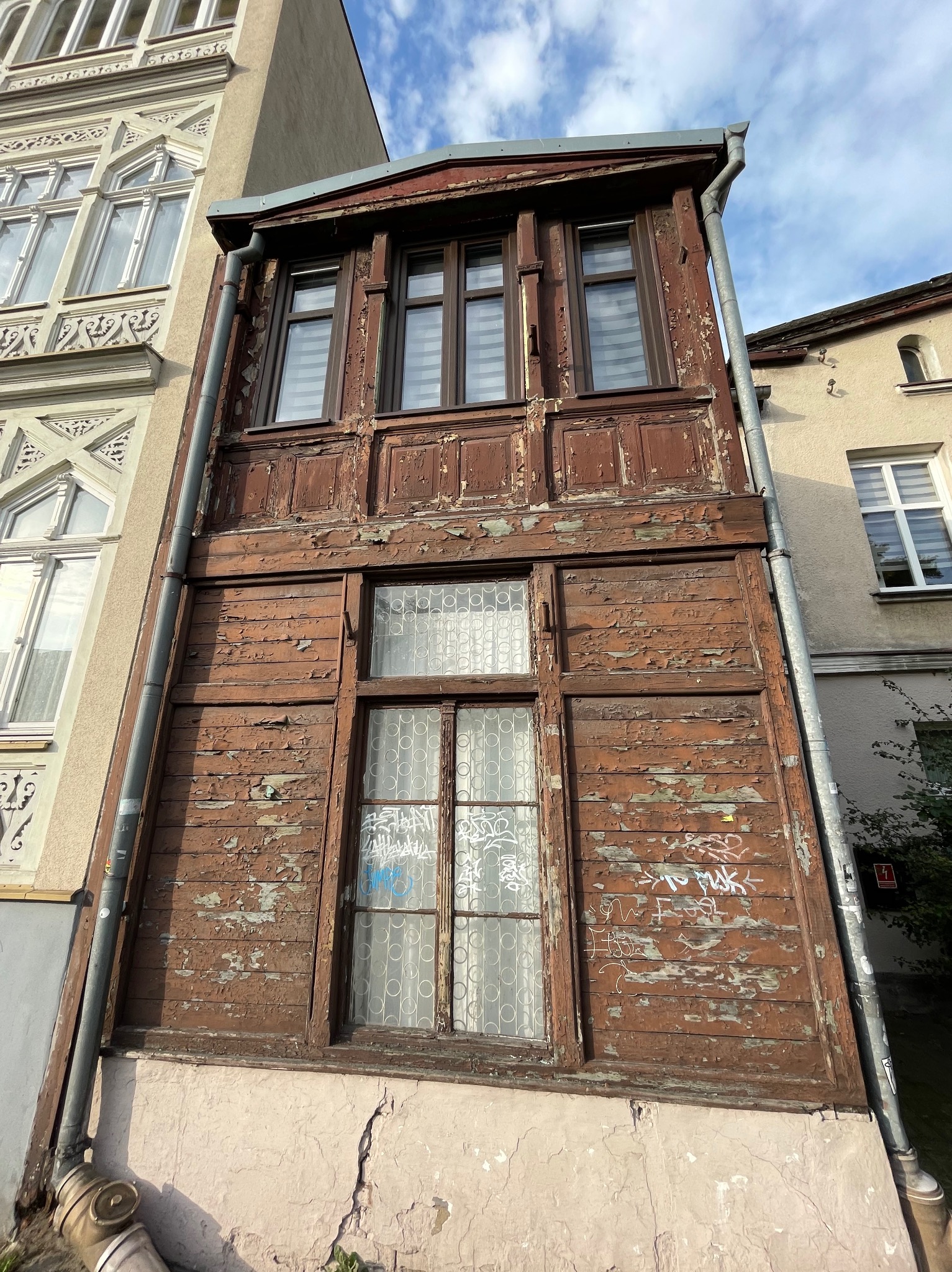 Stara, drewniana dobudówka do kamienicy z łuszczącą się farbą, graffiti i rynnami z boku, widok z dołu na tle błękitnego nieba.