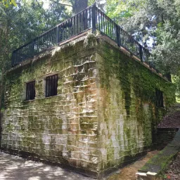 Kamienna altana z płaskim dachem i czarną metalową balustradą, porośnięta mchem, otoczona zielenią drzew.