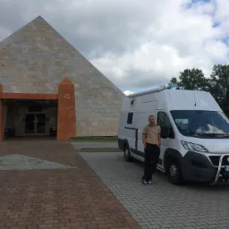 Mężczyzna oparty o biały samochód kempingowy na tle piramidy z kamienia, obok wejścia zdobionego motywami egipskimi.