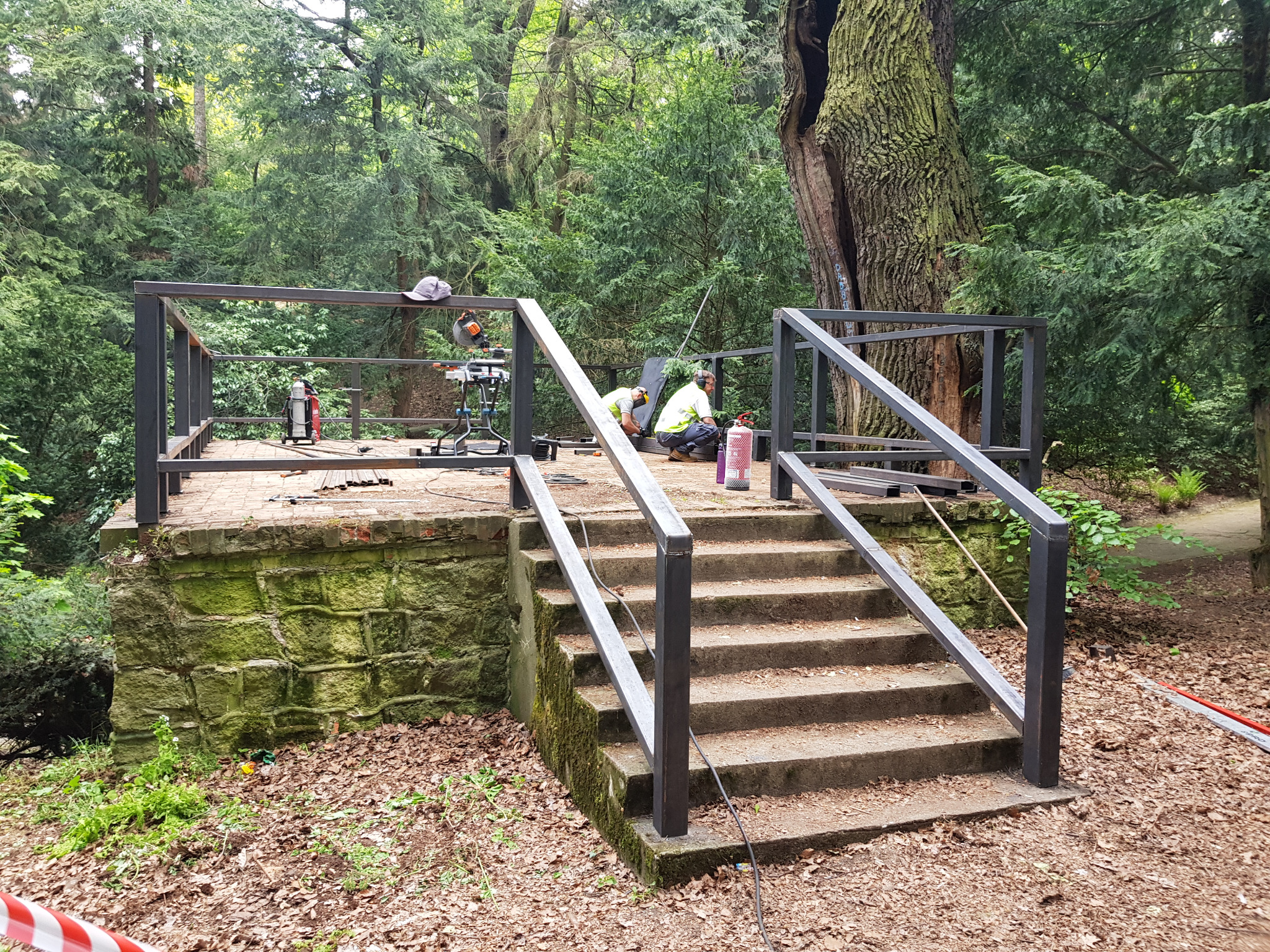 Montaż metalowej konstrukcji z poręczami na schodach w otoczeniu leśnym, dwóch pracowników w strojach roboczych, widoczne narzędzia i butla z gazem.
