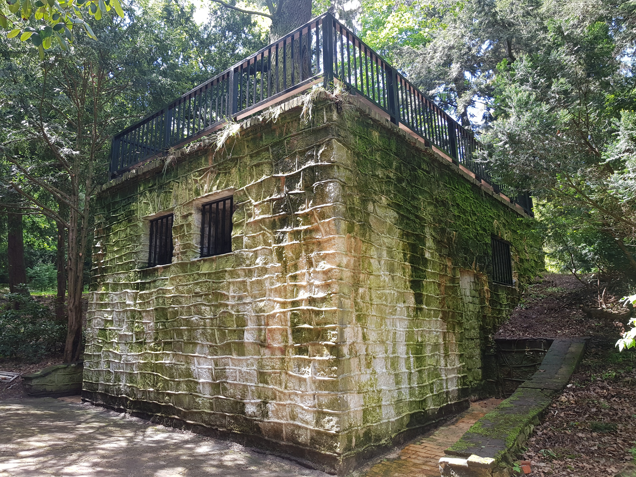 Kamienna altana z płaskim dachem i czarną metalową balustradą, porośnięta mchem, otoczona zielenią drzew.