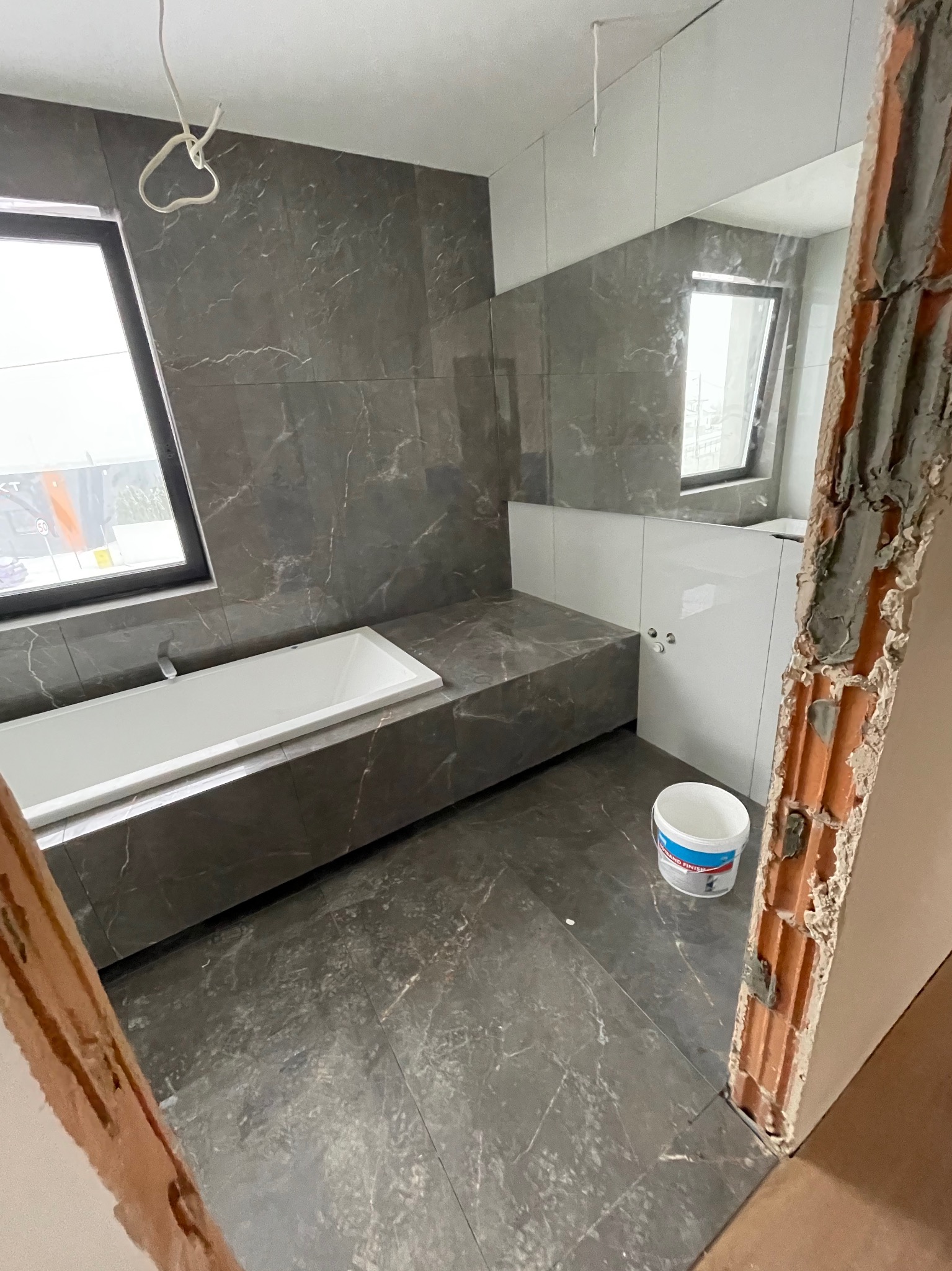 Łazienka w trakcie remontu: szare marmurowe płytki, biała wanna, widoczna puszka z farbą i surowe wykończenie ściany z cegły.