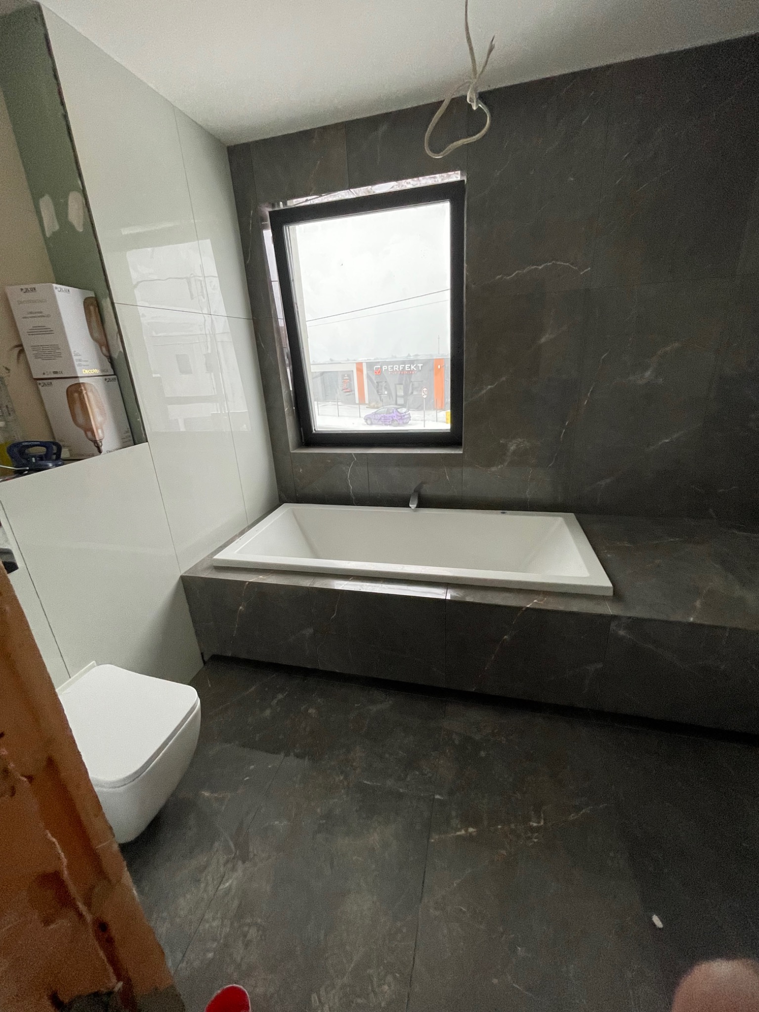Wanna zabudowana w ciemnych płytkach imitujących marmur pod oknem, biała toaleta w rogu, widoczne materiały budowlane, etap prac wykończeniowych.