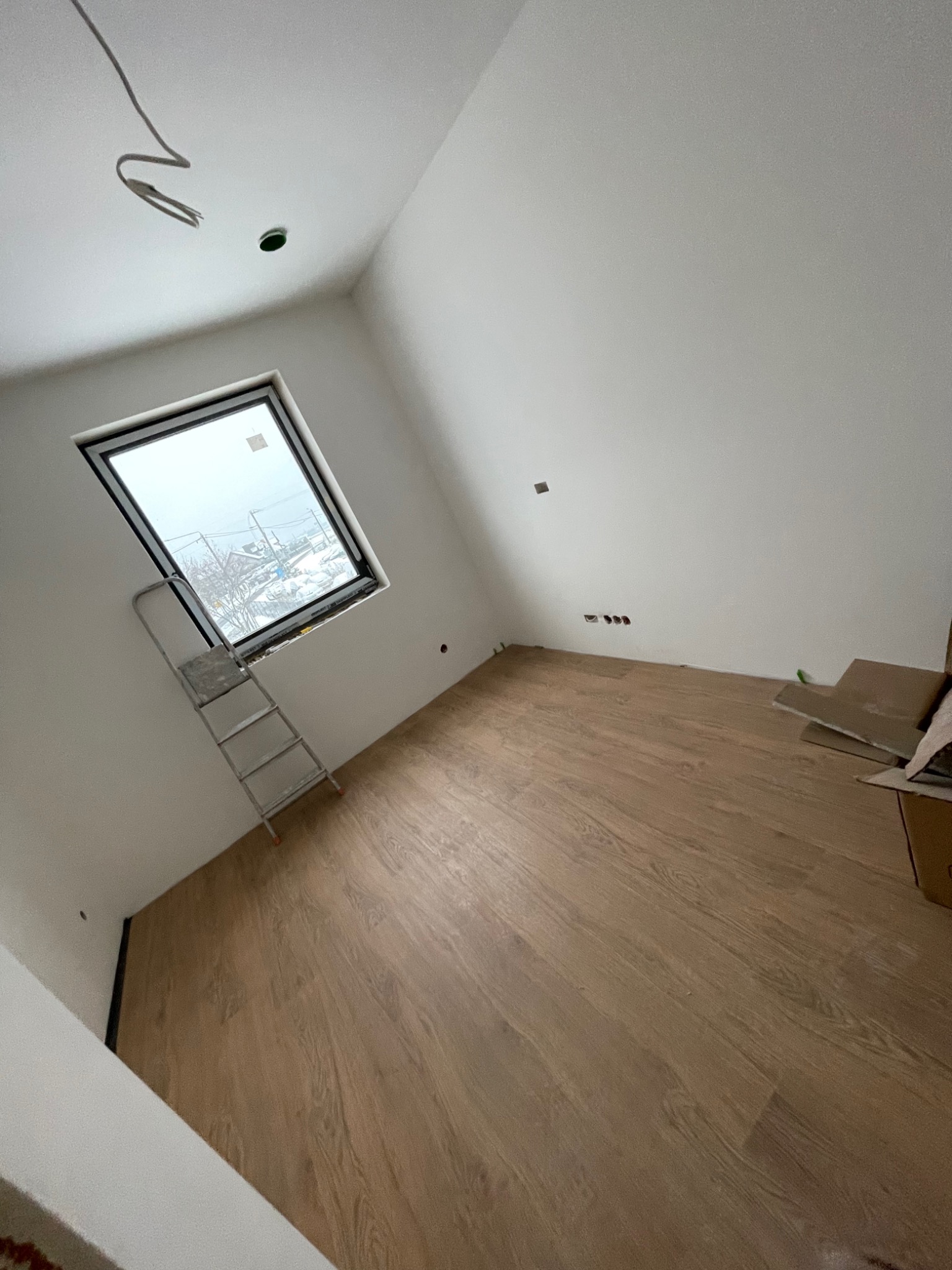 Poddasze z nową drewnianą podłogą, drabina oparta o okno dachowe, widoczne gniazdka elektryczne i wystające kable z sufitu.
