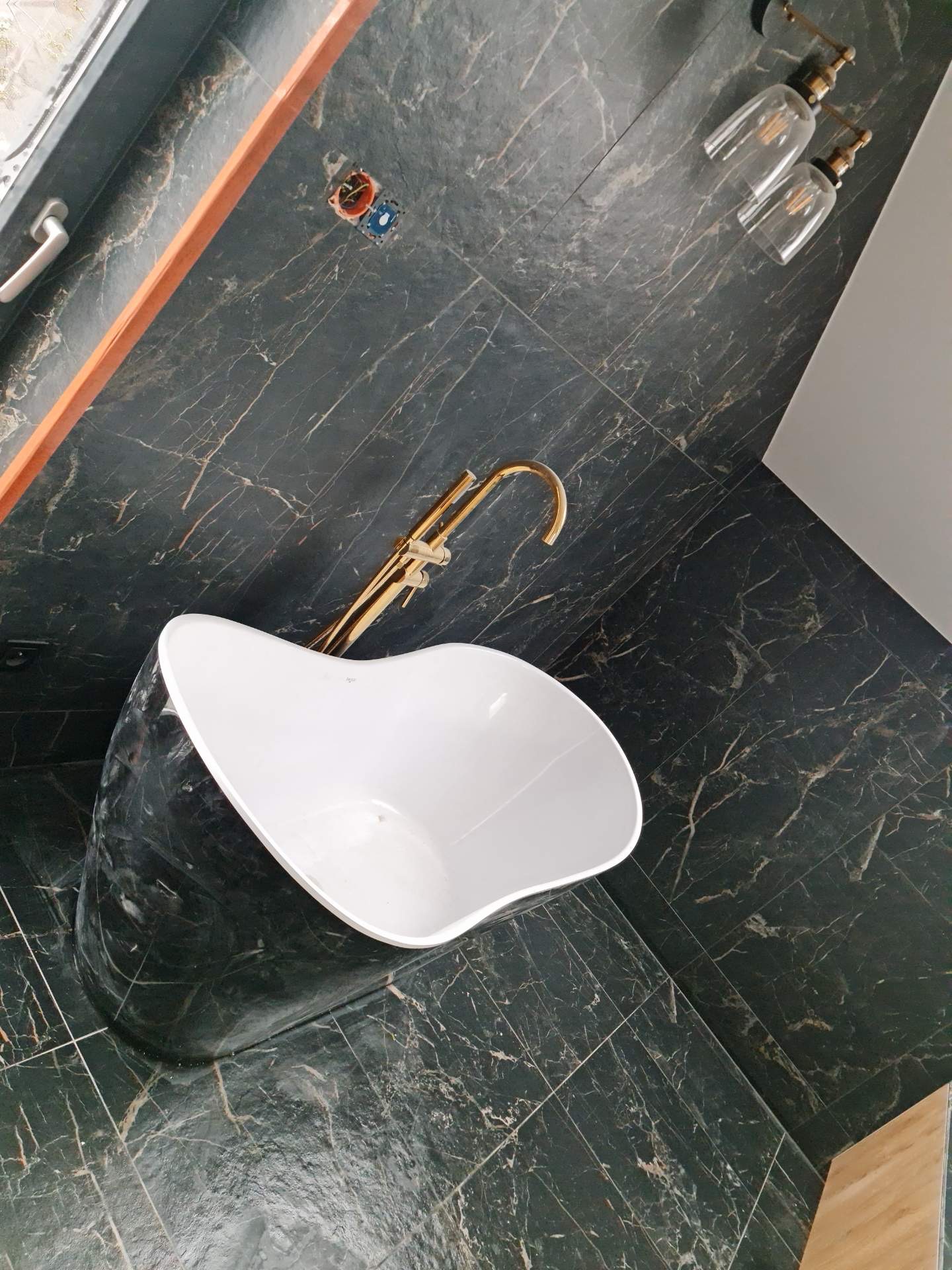 Elegancka wolnostojąca wanna w łazience wyłożonej ciemnym marmurem, z baterią w kolorze złota i designerskimi lampami sufitowymi.