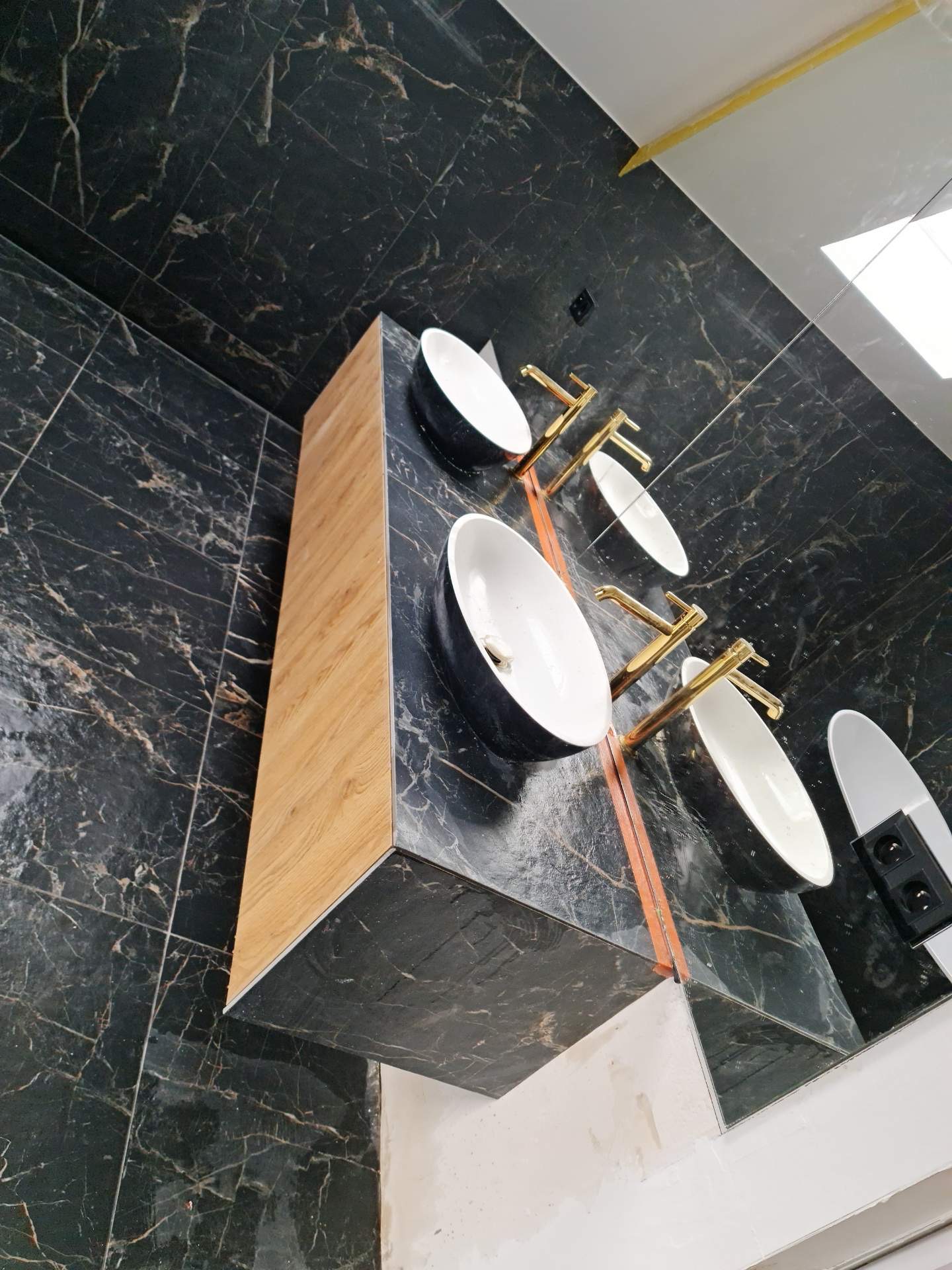 Nowoczesna łazienka z trzema umywalkami nablatowymi w czarnych obudowach i złotymi bateriami, zamontowanymi na blacie z drewnopodobnym wykończeniem, ściany i podłoga wyłożone czarnymi płytkami...