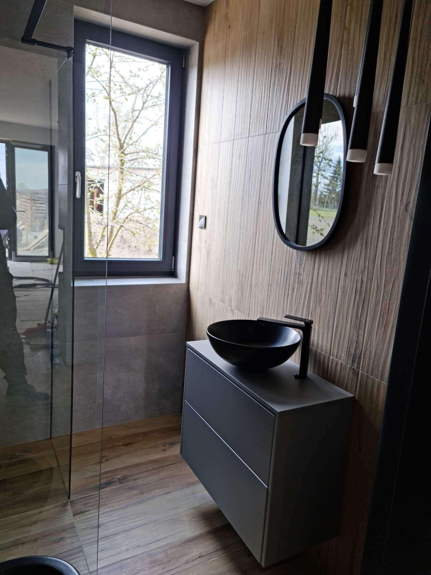 Nowoczesna łazienka z drewnopodobnymi płytkami ściennymi, czarną umywalką nablatową i minimalistyczną szafką w kolorze grafitowym, oświetlona wiszącymi lampami tubami.
