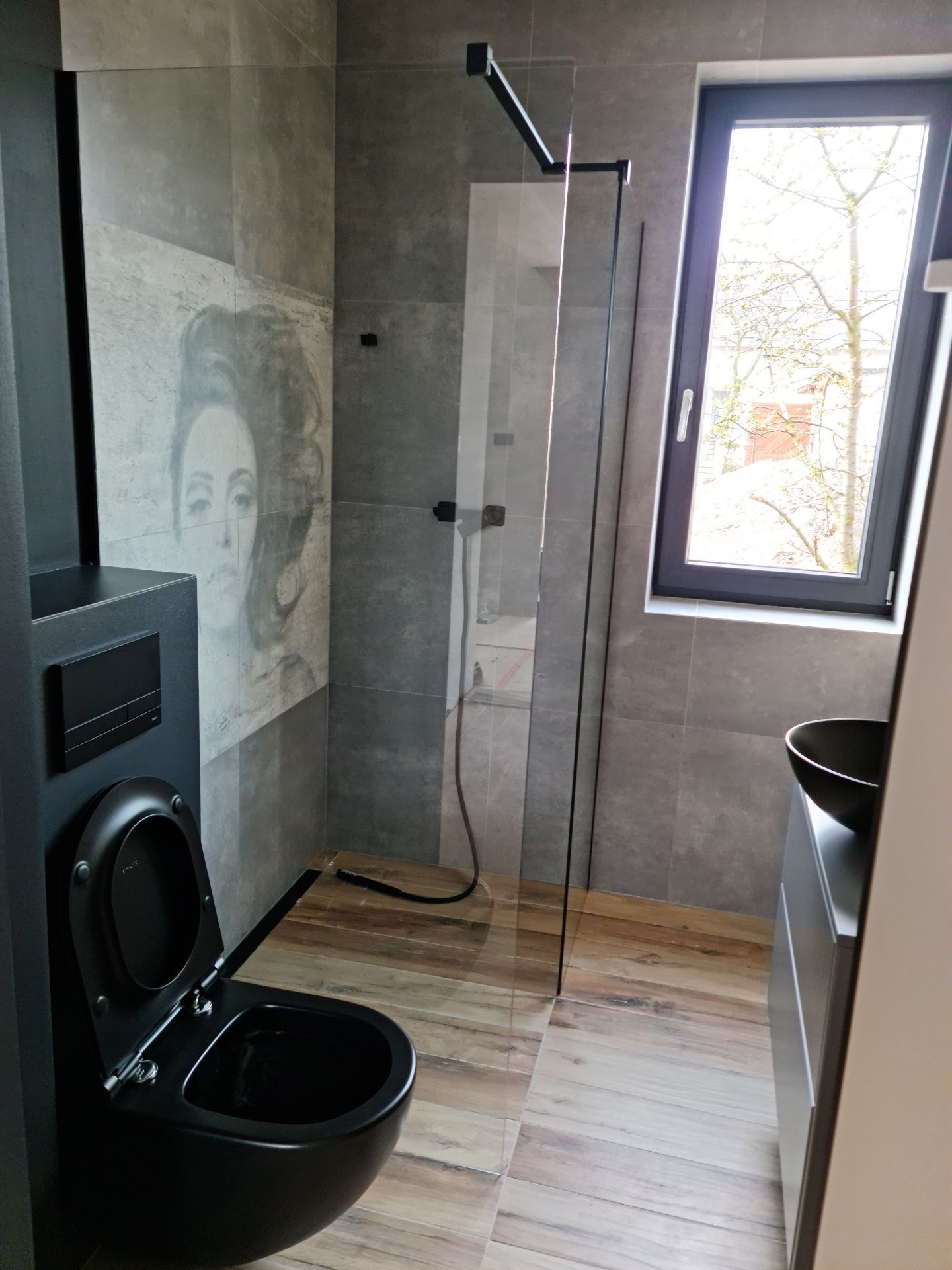 Nowoczesna łazienka z szarymi płytkami, drewnopodobną podłogą, czarną armaturą i kabiną prysznicową typu walk-in, wyróżniająca się portretem kobiety na ścianie.