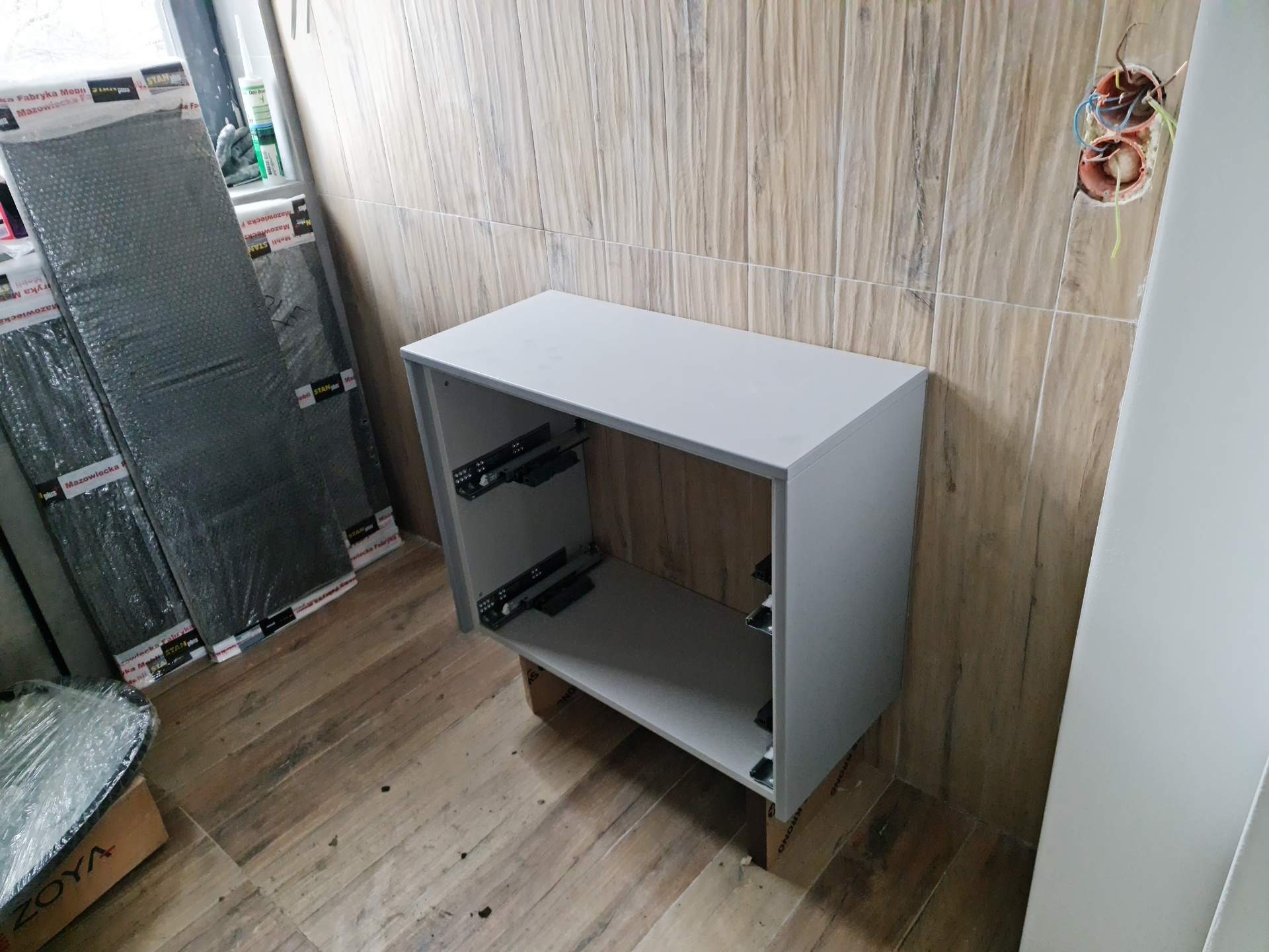 Montaż szarej szafki pod umywalkę w łazience z beżowymi płytkami imitującymi drewno, widoczne niezabezpieczone kable elektryczne w ścianie, elementy montażowe i opakowania po produktach.