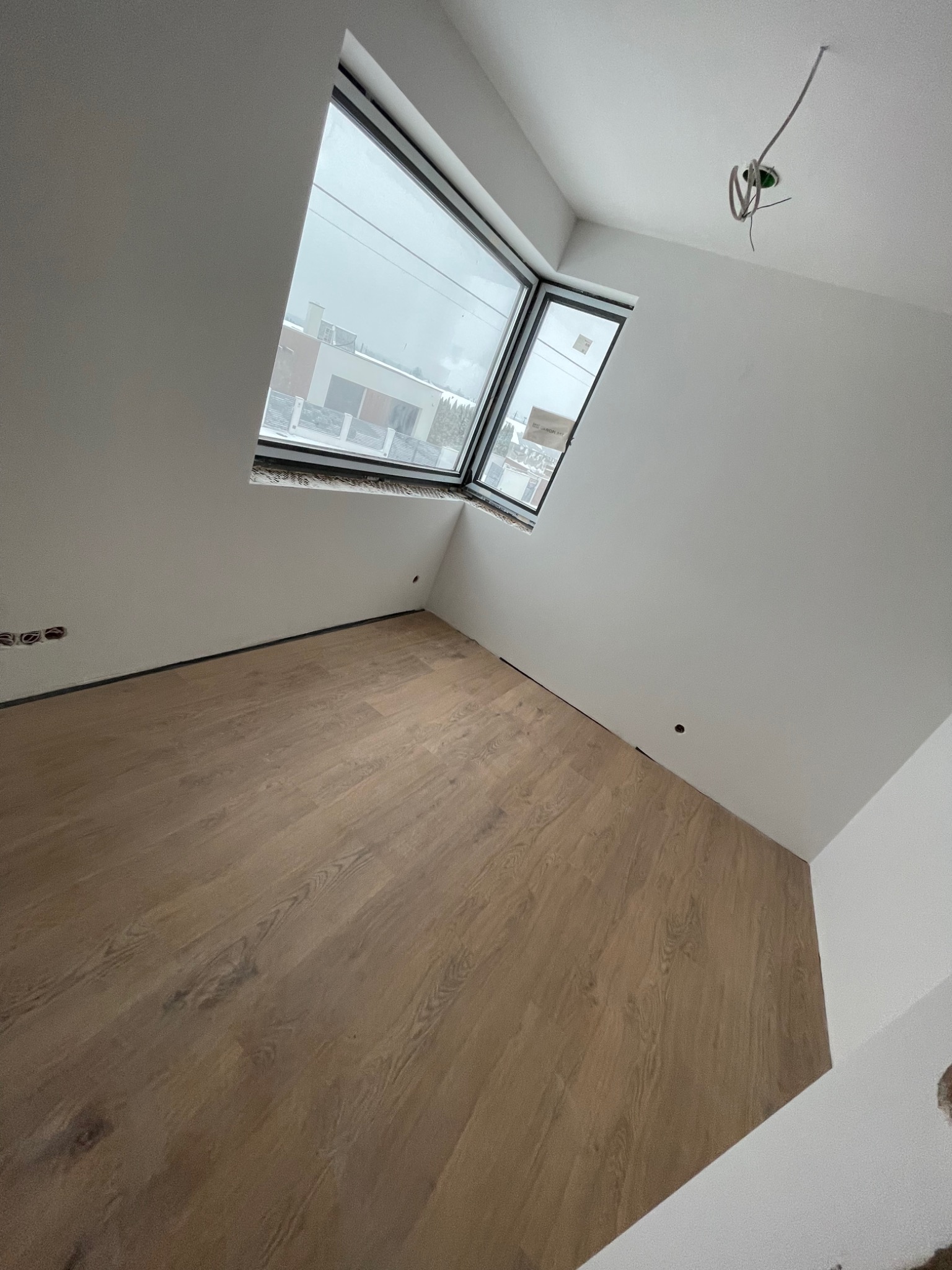 Pokój po remoncie z drewnianą podłogą, białymi ścianami i oknem z widokiem na budynek z zewnątrz. Widoczne wystające przewody elektryczne na suficie.