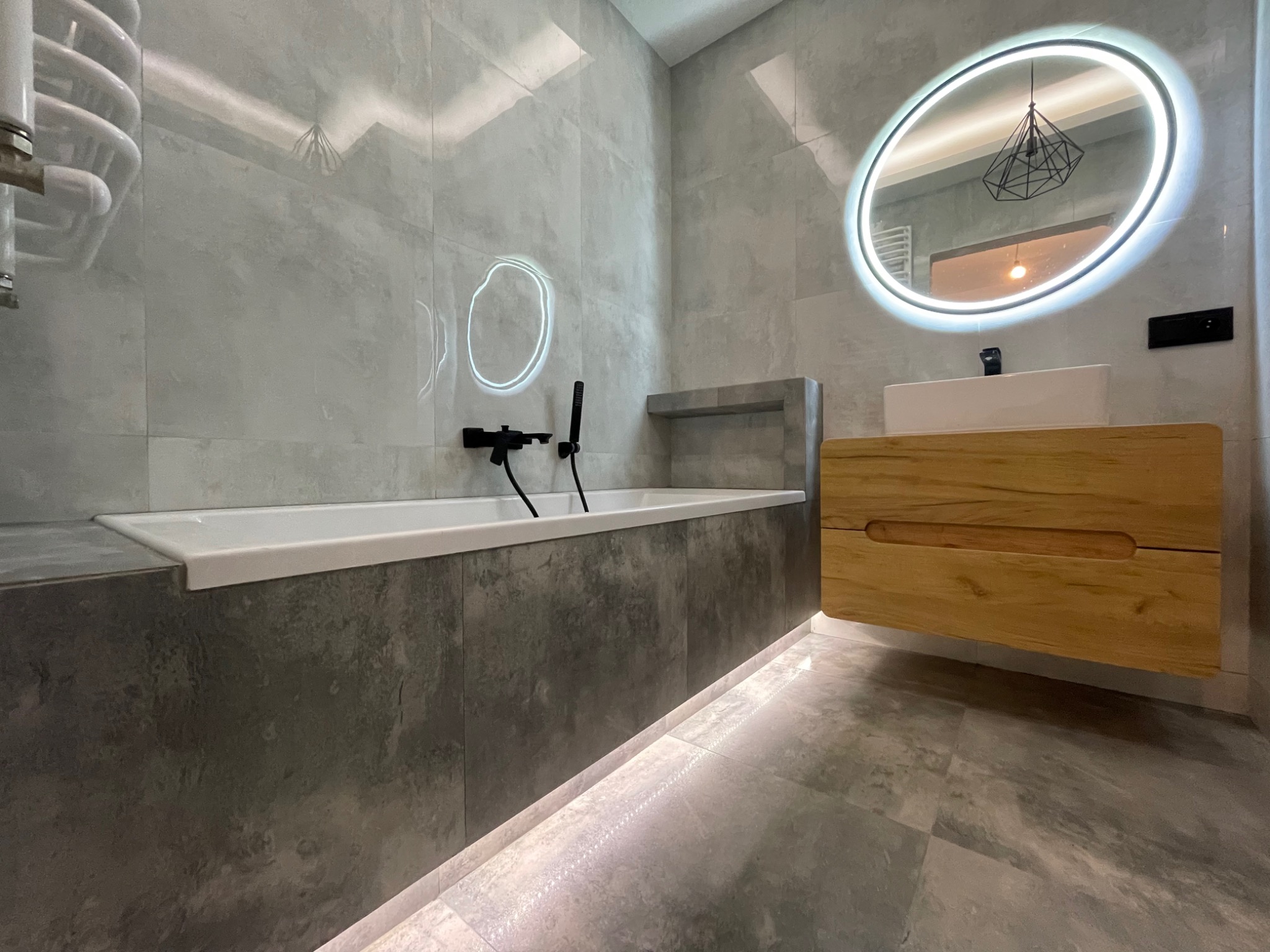 Nowoczesna łazienka z szarymi płytkami imitującymi beton, podświetlaną wanną, drewnianą szafką pod umywalką i okrągłym lustrem z podświetleniem LED oraz czarną armaturą.