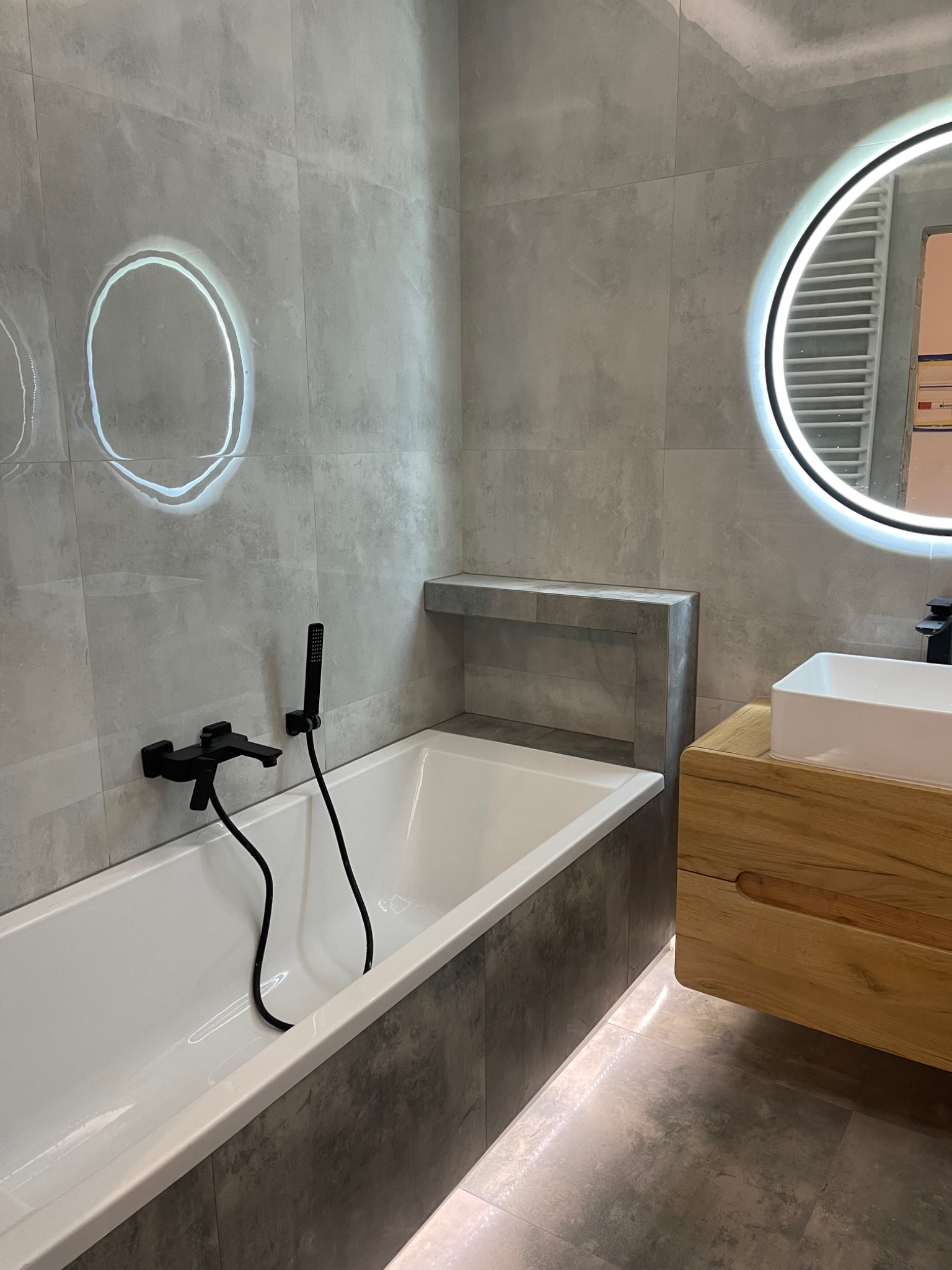 Nowoczesna łazienka z szarymi płytkami, białą wanną z czarną armaturą, drewnianą szafką pod umywalką i okrągłym lustrem z podświetleniem LED.