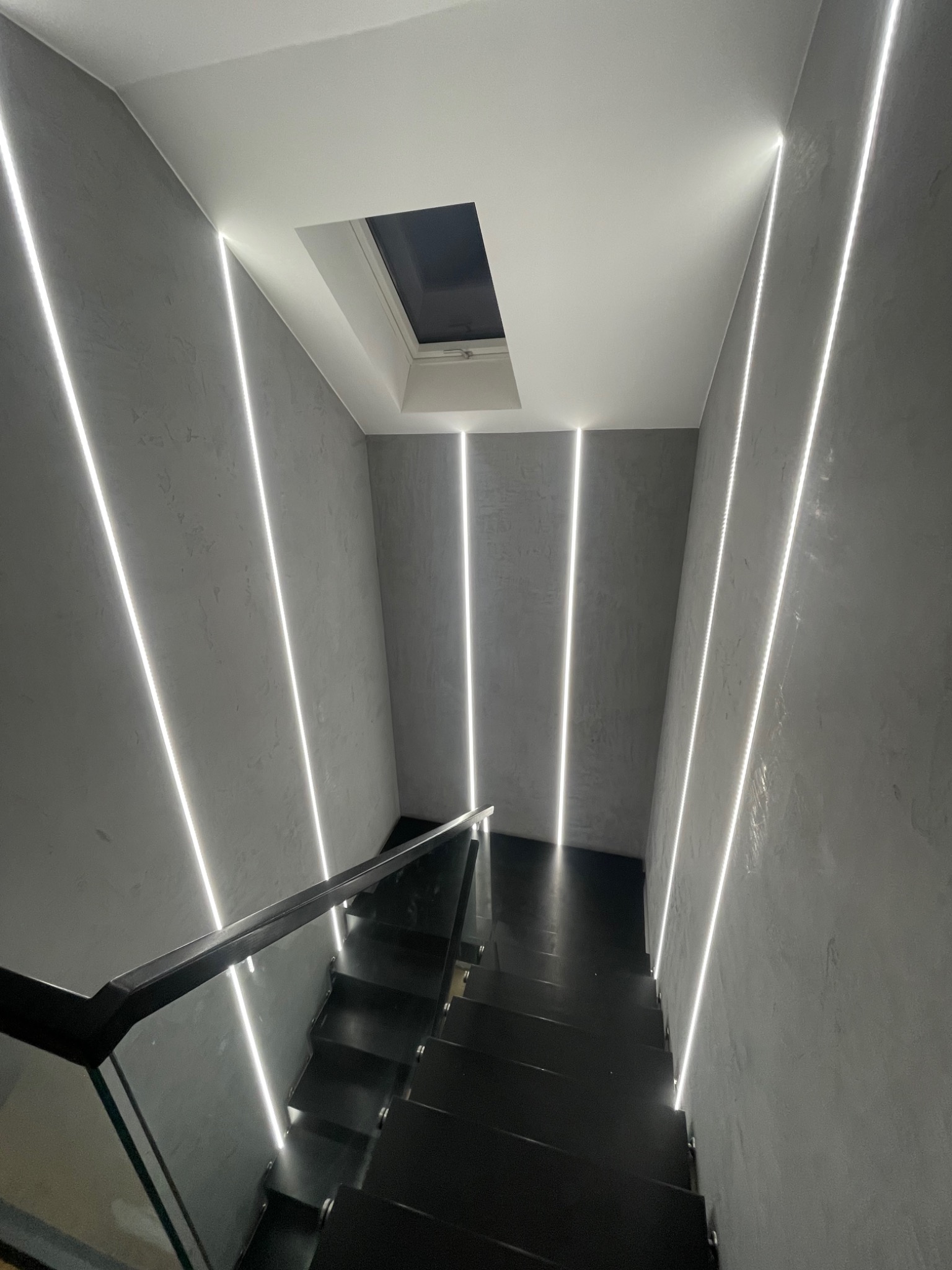 Nowoczesne schody z czarnymi stopniami i szklaną balustradą, oświetlone pionowymi pasami LED na szarych ścianach o fakturze betonu, z widocznym oknem dachowym.