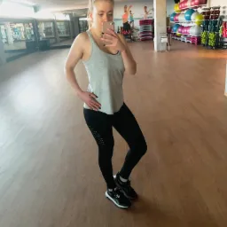 Kobieta w sportowym stroju robi selfie w lustrze na siłowni, w tle widoczne piłki do ćwiczeń i grafika na ścianie.