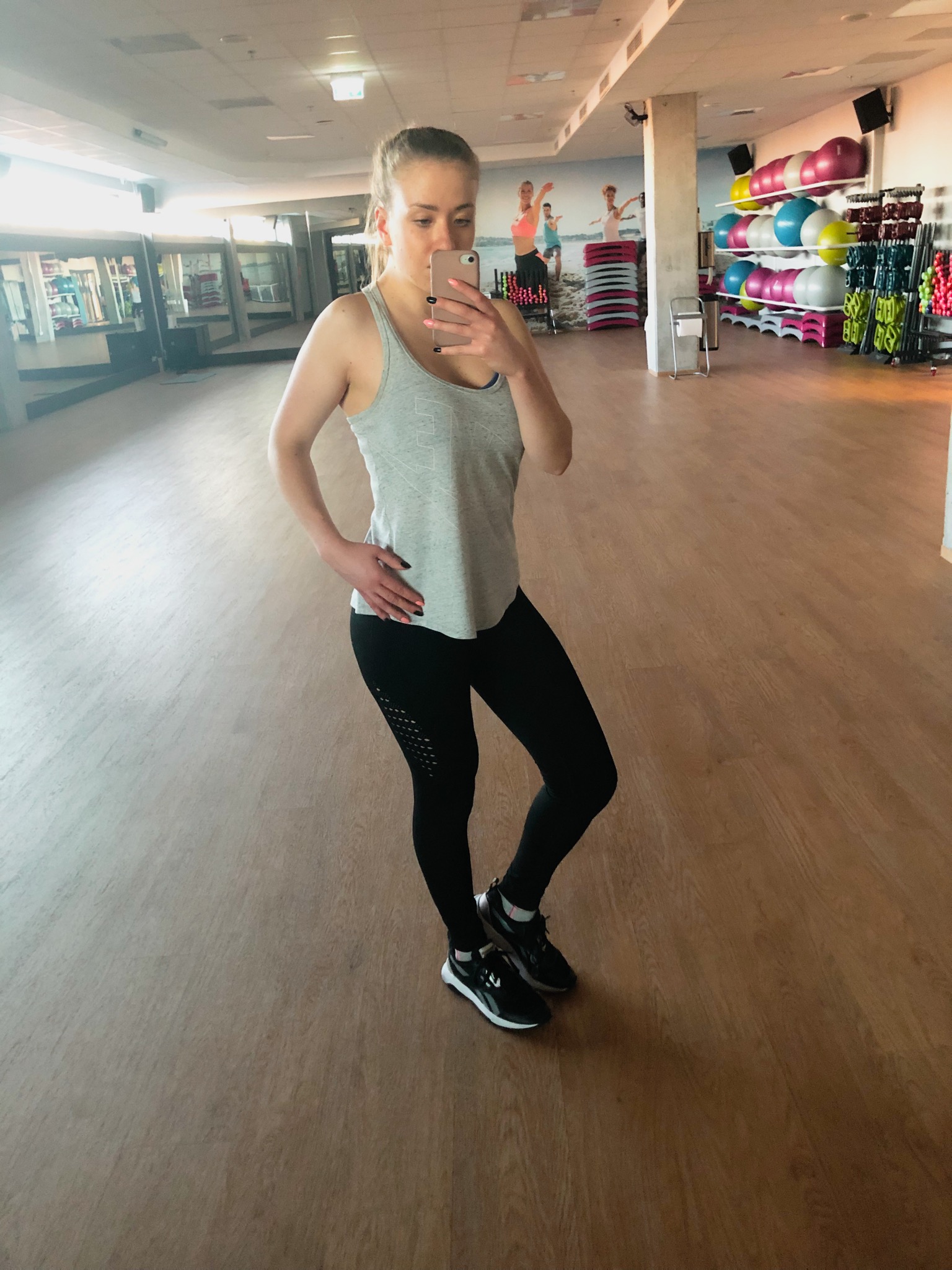 Kobieta w sportowym stroju robi selfie w lustrze na siłowni, w tle widoczne piłki do ćwiczeń i grafika na ścianie.