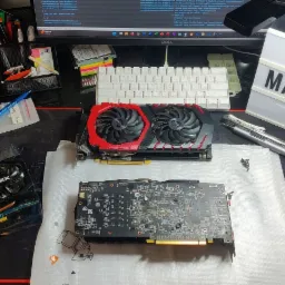 Trzy karty graficzne na macie z mapą świata, narzędzia i śrubki obok, podświetlana tablica z napisem 'MAC IT' w tle, monitor Dell wyświetlający kod.