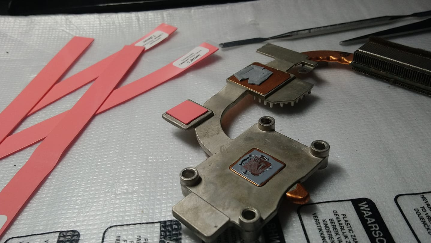 Metalowy radiator z pozostałościami pasty termoprzewodzącej i nowymi termopadami, przygotowany do montażu w laptopie, obok leżą narzędzia i etykiety z grubością termopadów