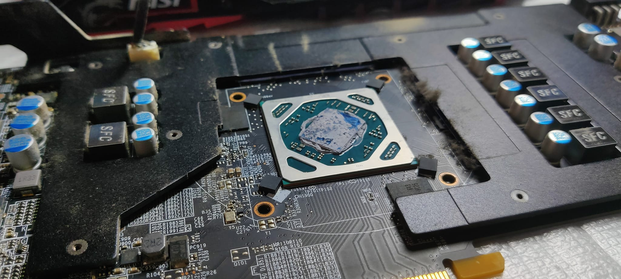 Otwarta karta graficzna MSI z widocznym procesorem graficznym pokrytym pastą termoprzewodzącą przed czyszczeniem, widoczne elementy elektroniki i ślady kurzu.