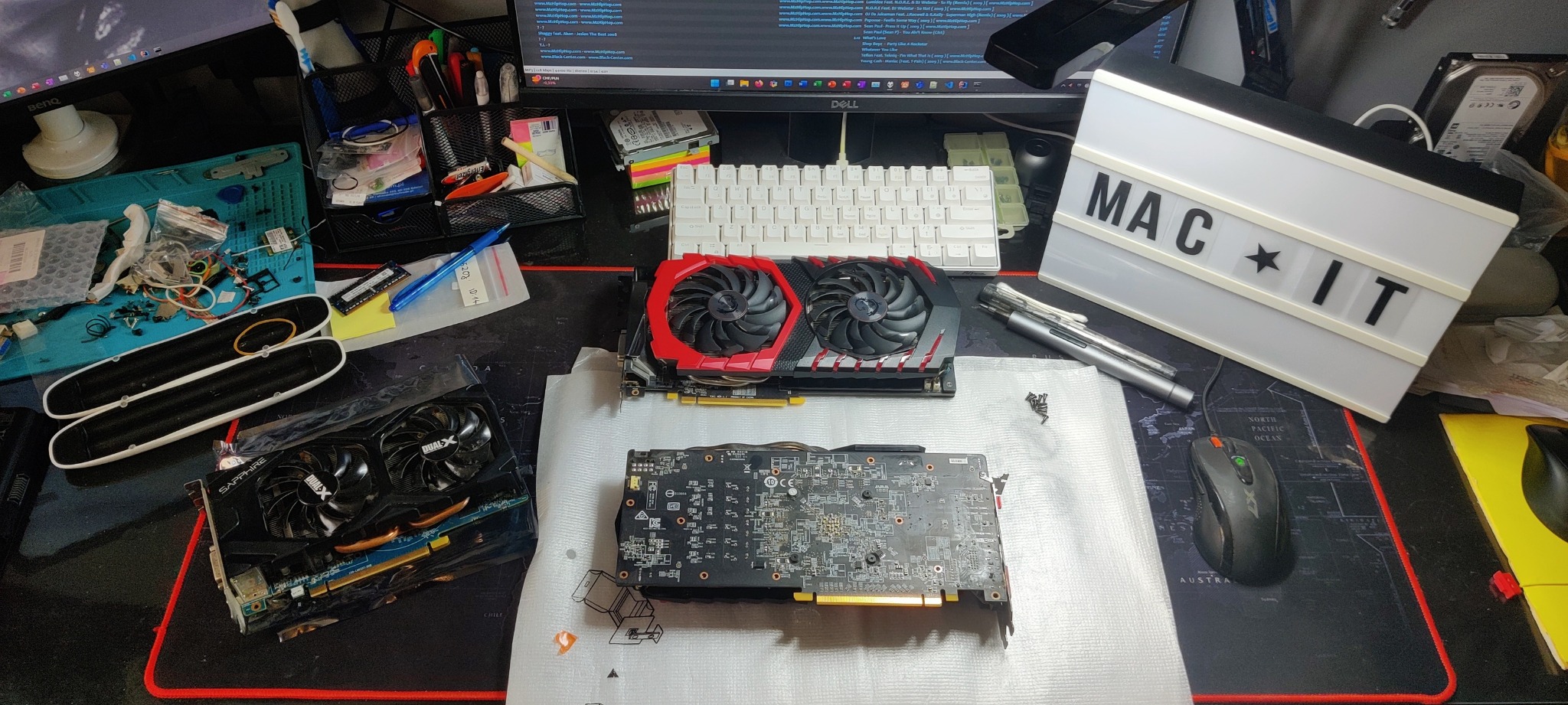 Trzy karty graficzne na macie z mapą świata, narzędzia i śrubki obok, podświetlana tablica z napisem 'MAC IT' w tle, monitor Dell wyświetlający kod.