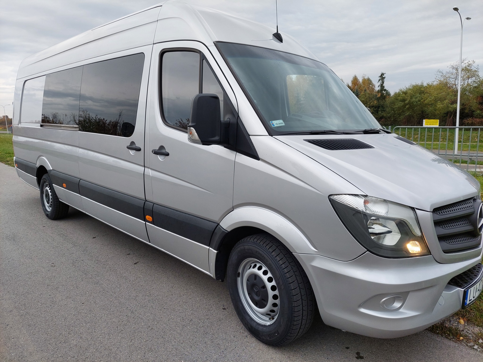Srebrny bus Mercedes-Benz Sprinter z przyciemnianymi szybami, zaparkowany na asfaltowej drodze, widok z przodu i boku, z widoczną tablicą rejestracyjną.