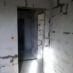 Wnętrze w stanie surowym, widoczne białe ściany z betonu komórkowego, rozprowadzone przewody elektryczne w peszlach oraz puszki instalacyjne. Na ścianie widoczne notatki ołówkiem.