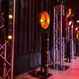 Rząd stalowych konstrukcji kratownicowych i lamp oświetleniowych w kolorach żółtym i czerwonym, ustawionych na scenie z czarną kotarą w tle.