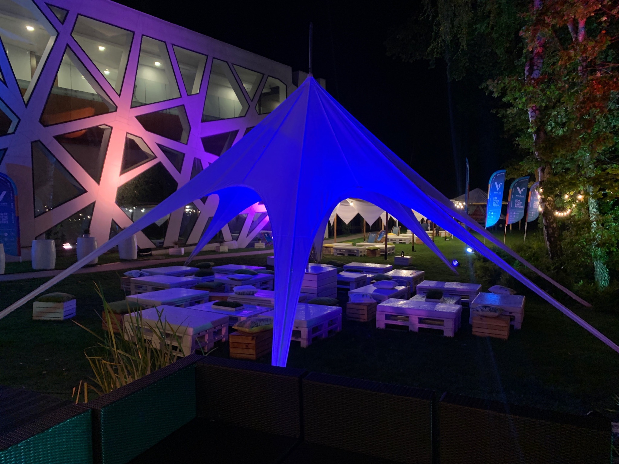 Oświetlona, niebieska konstrukcja namiotowa na trawie, z paletowymi stolikami i siedziskami, budynek w tle oświetlony na różowo, wieczorna sceneria.