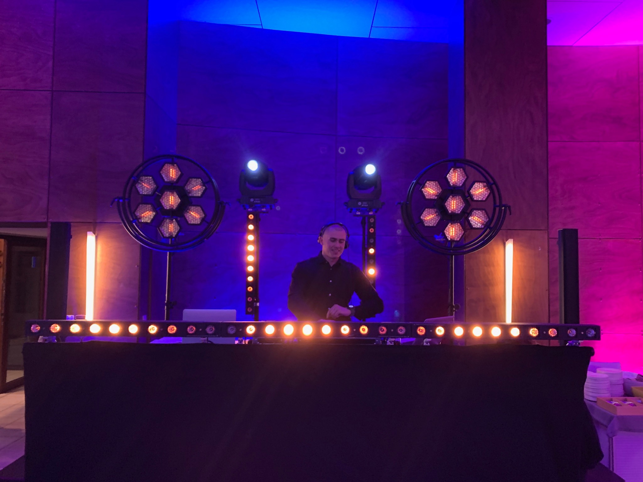 DJ uśmiecha się za konsoletą oświetloną ciepłym światłem, z efektami świetlnymi w tle, w sali z niebiesko-różowym oświetleniem.