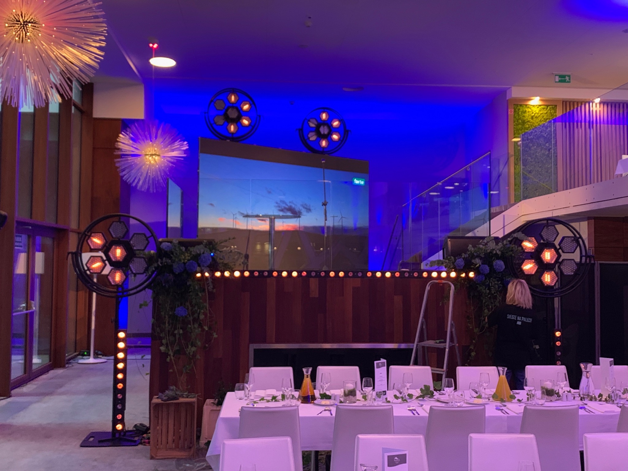 Uroczysta sala bankietowa oświetlona niebieskim światłem z dekoracjami kwiatowymi i stołami przygotowanymi do przyjęcia, z widocznymi reflektorami i ekranem wyświetlającym zachód słońca.