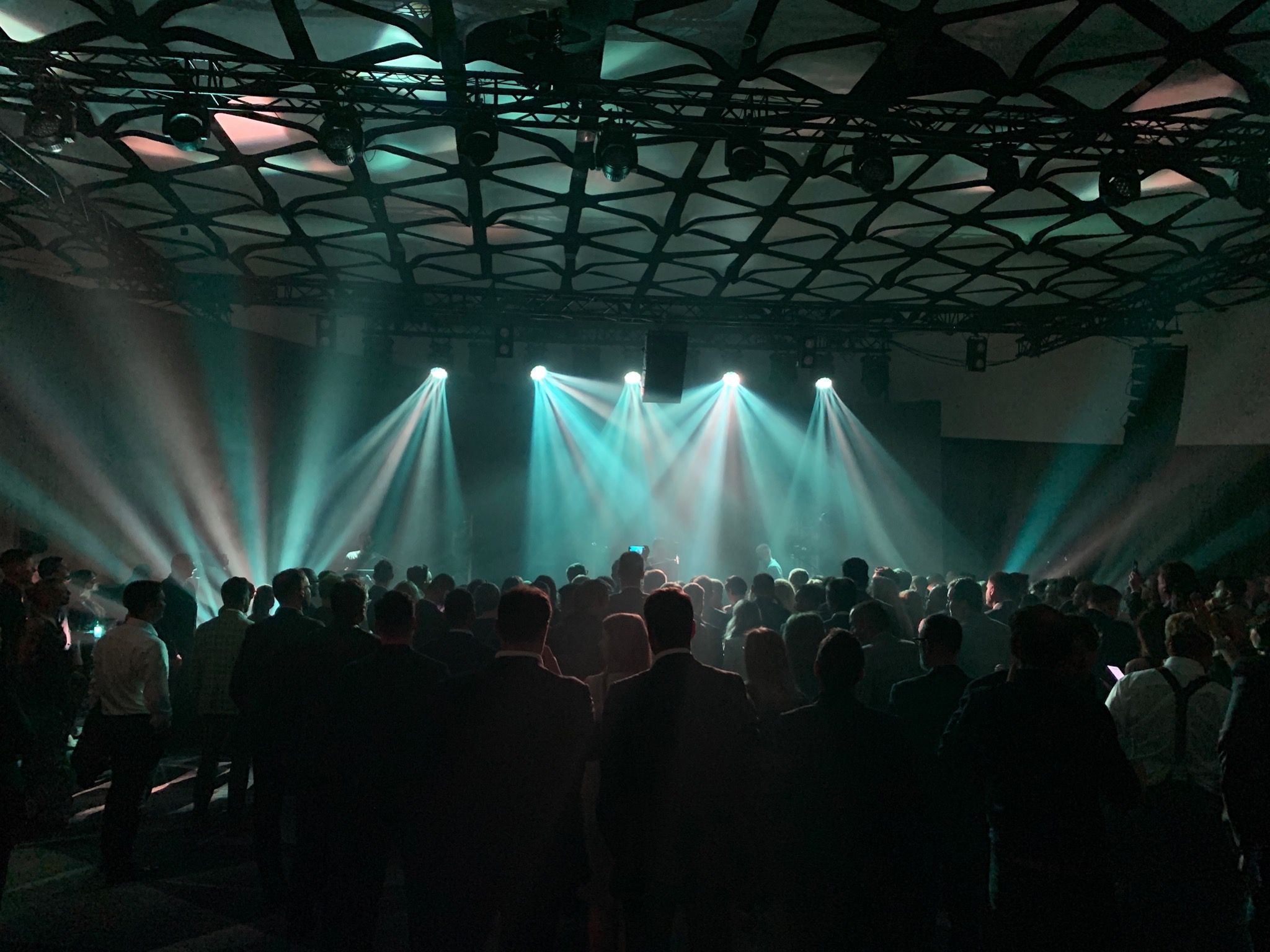 Tłum ludzi na widowni podczas wydarzenia oświetlonego reflektorami z zielonkawą poświatą, widoczna konstrukcja sufitu i głośniki sceniczne.
