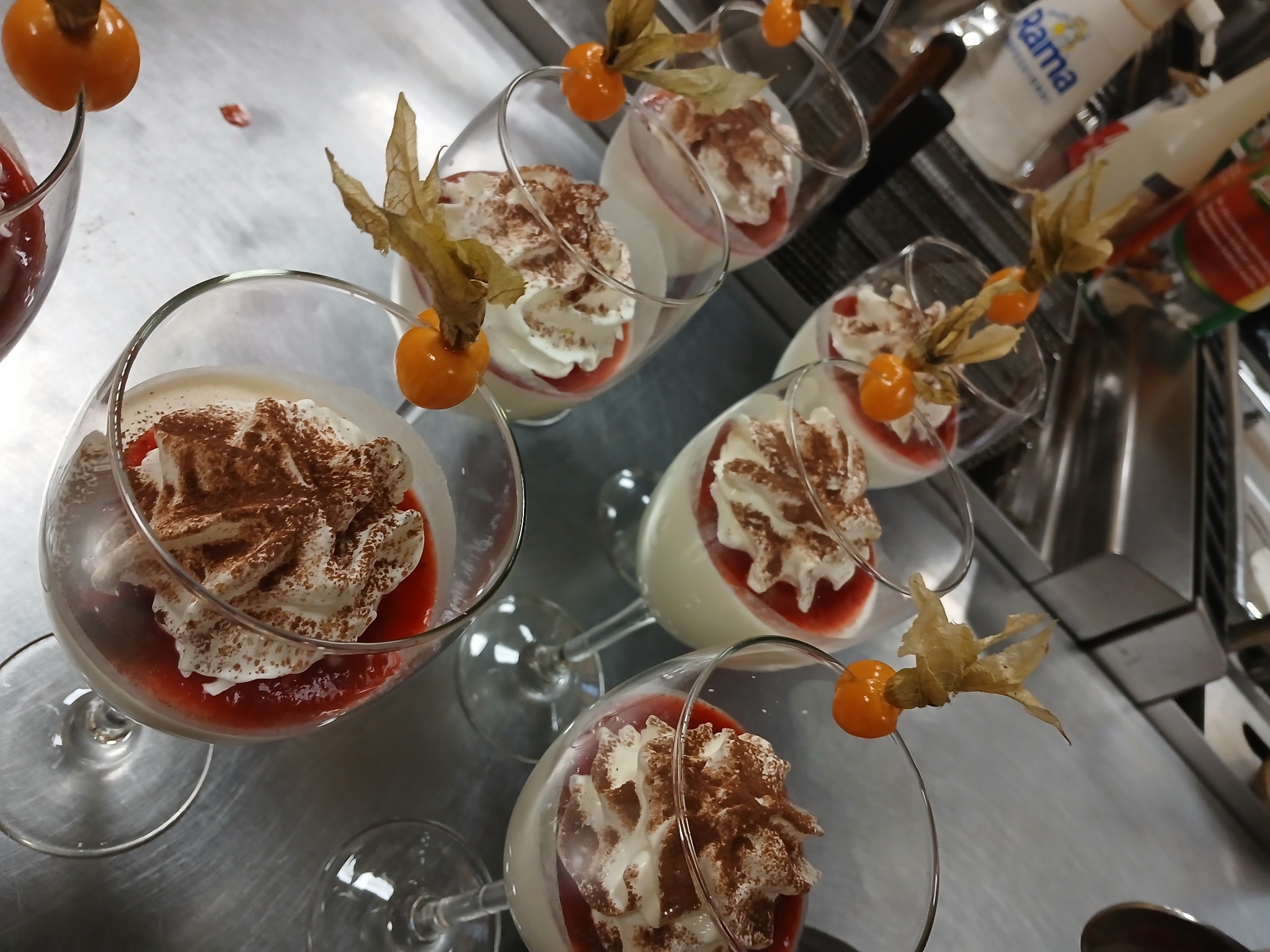Eleganckie desery w szklankach, udekorowane bitą śmietaną, kakao i physalis, ustawione na metalowym blacie, gotowe do podania. Widok z góry.