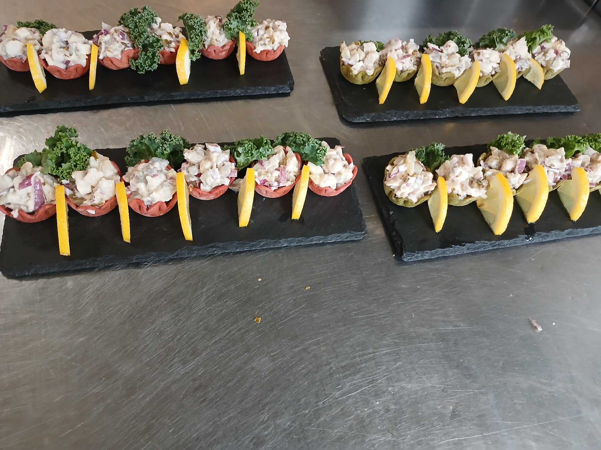 Elegancko podane przystawki na czarnych deskach: sałatka w czerwonych i zielonych babeczkach, udekorowana cytryną i zielonymi liśćmi, na stalowym blacie.