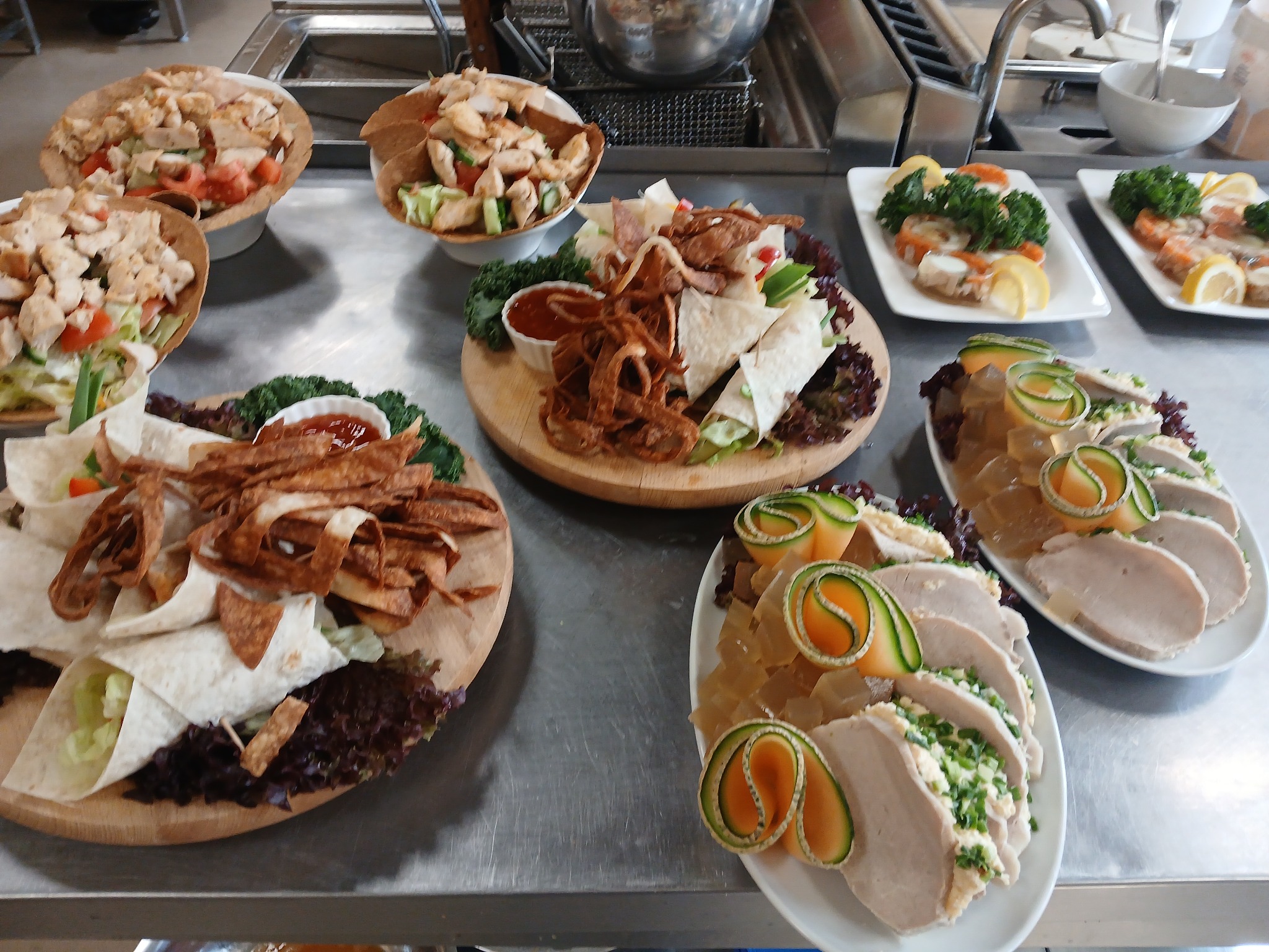 Profesjonalnie przygotowane, różnorodne przekąski cateringowe: tortille, sałatki, mięsa, dekorowane warzywami, ułożone na talerzach i deskach. Widok z góry na metalowym blacie.