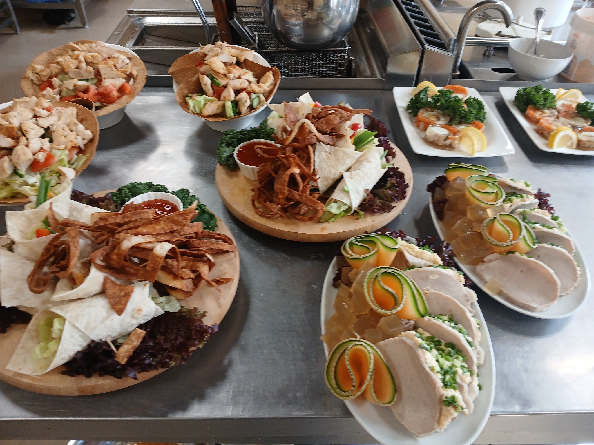 Różnorodne dania cateringowe: sałatki w tortillach, roladki z szynki z melonem i galaretką, przystawki z krewetkami i cytryną, na metalowym blacie kuchennym.