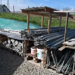 Skladowane stemple budowlane w drewnianych regałach pod zadaszeniem, widoczne dwa białe wiadra z farbą, w tle dom i zielona siatka zabezpieczająca.