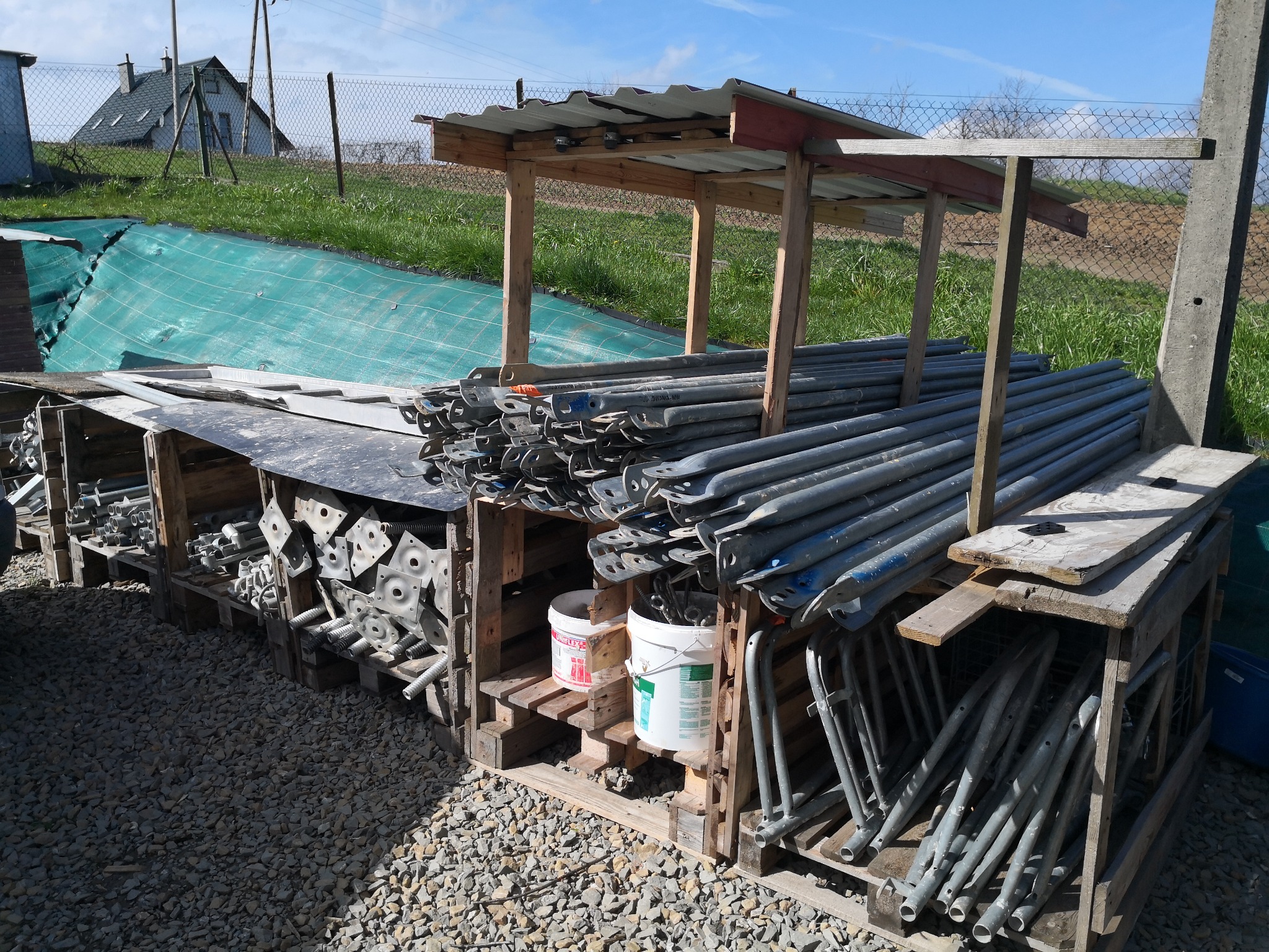 Skladowane stemple budowlane w drewnianych regałach pod zadaszeniem, widoczne dwa białe wiadra z farbą, w tle dom i zielona siatka zabezpieczająca.