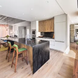 Jasny parkiet ułożony w jodełkę francuską w nowoczesnej kuchni z wyspą i zielonymi hokerami, widok na jadalnię i okno z widokiem na miasto.