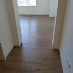 Ułożona podłoga z paneli w odcieniach brązu, widok z perspektywy wejścia do pomieszczenia, białe ściany i listwy przypodłogowe, okno w tle.