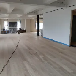 Jasna drewniana podłoga w przestronnym wnętrzu biurowym po cyklinowaniu, z widocznymi śladami po maszynie i zabezpieczonymi folią oknami i meblami, czarna kolumna na środku pomieszczenia.