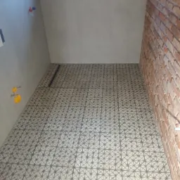 Wykończona łazienka z wzorzystą, szarą podłogą z odpływem liniowym, jasnymi płytkami ściennymi i ścianą z cegły. Widoczne elementy instalacji wodnej.